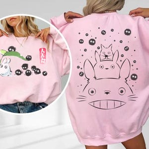 May include: A light pink sweatshirt featuring a cartoon design. The front has a white rabbit-like character with black soot sprites and Japanese text. The back displays a larger graphic of the same characters. A comfortable, casual style.
