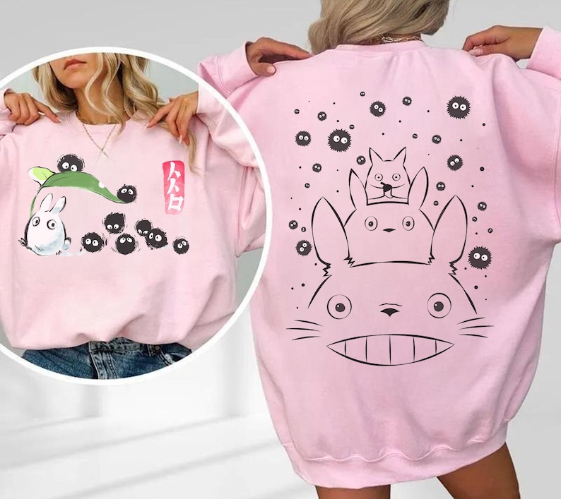 May include: A light pink sweatshirt featuring a cartoon design. The front has a white rabbit-like character with black soot sprites and Japanese text. The back displays a larger graphic of the same characters. A comfortable, casual style.