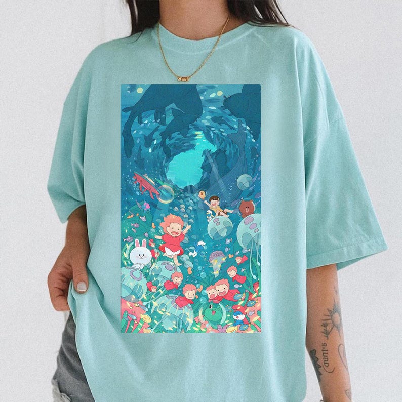 K&ouml;nnte beinhalten: Hellblaues T-Shirt mit einer farbenfrohen Illustration einer Unterwasserszene mit Zeichentrickfiguren, Fischen und Blasen. Das grafische Design ist mittig platziert und zeigt eine lebendige, skurrile Unterwasserwelt.