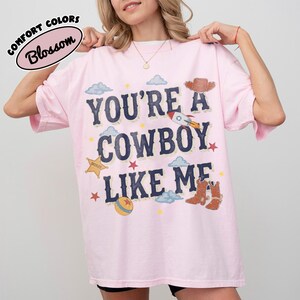 K&ouml;nnte beinhalten: Ein hellrosa T-Shirt mit dem dunkelblauen Schriftzug "YOU'RE A COWBOY LIKE ME" und grafischen Illustrationen einer Rakete, eines Sheriffsterns, eines Balls, Cowboystiefeln, einem Cowboyhut, Wolken und kleinen Sternen auf der Vorderseite.