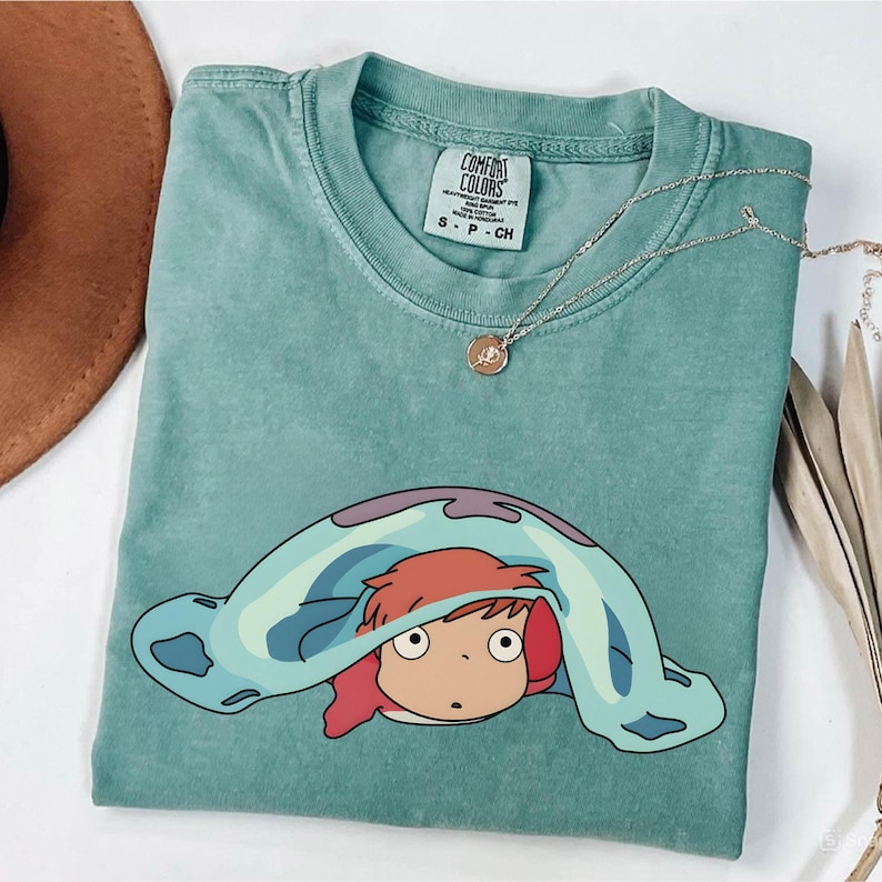 May include: A teal Comfort Colors t-shirt featuring a cartoon character with red hair and a blue blanket. The shirt has a crew neck and short sleeves. A gold necklace with a pendant is draped across the shirt.