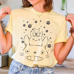 May include: A light yellow t-shirt featuring a black and white graphic of three cartoon characters. The largest character has a wide grin, and the other two are stacked on top. Small black dots are scattered around the characters.