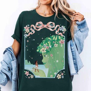 May include: Dark green t-shirt featuring a framed illustration of a green-skinned woman with a floral crown and a woman with a dog on a beach. The design includes a pink ribbon and floral accents. The shirt is a casual wear item.