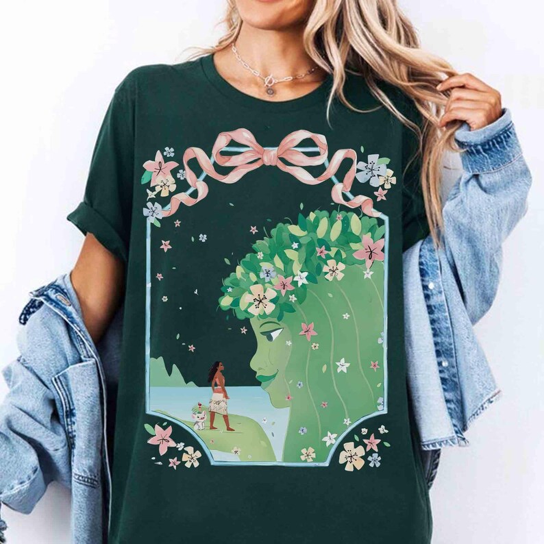 May include: Dark green t-shirt featuring a framed illustration of a green-skinned woman with a floral crown and a woman with a dog on a beach. The design includes a pink ribbon and floral accents. The shirt is a casual wear item.