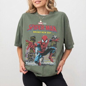 May include: Olive green t-shirt featuring a Spider-Man graphic. The graphic includes the words "Marvel Studios Spider-Man Brand New Day" with an illustration of Spider-Man in a city setting. The shirt has short sleeves and a crew neck.