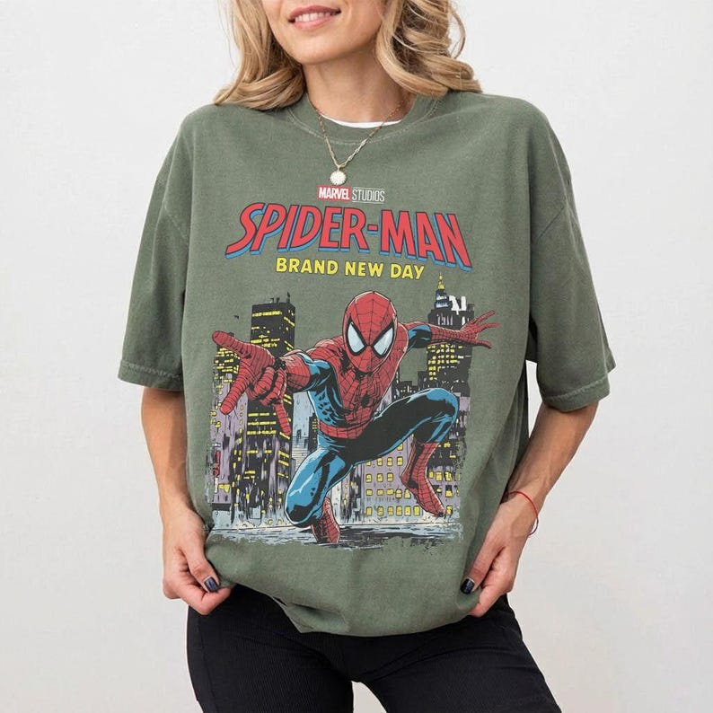 May include: Olive green t-shirt featuring a Spider-Man graphic. The graphic includes the words "Marvel Studios Spider-Man Brand New Day" with an illustration of Spider-Man in a city setting. The shirt has short sleeves and a crew neck.