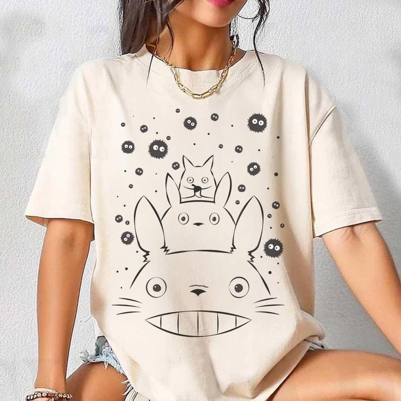 May include: Cream-colored t-shirt featuring a black and white graphic of a large, smiling cartoon character with two smaller characters on top, and several small, round, black figures scattered around the design.