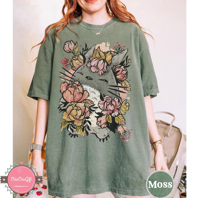 May include: Moss green t-shirt featuring a graphic of a gray cartoon cat surrounded by pink and yellow flowers. The cat has a white belly and is smiling. The shirt is a comfortable, oversized fit.