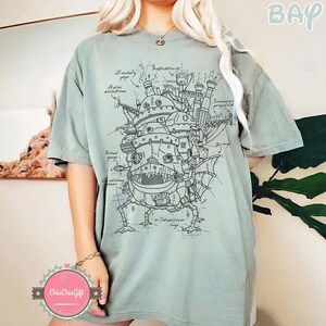 May include: Sage green t-shirt featuring a detailed black and white illustration of a fantastical, mechanical structure with architectural details and labeled components. The design is reminiscent of a blueprint or technical drawing.