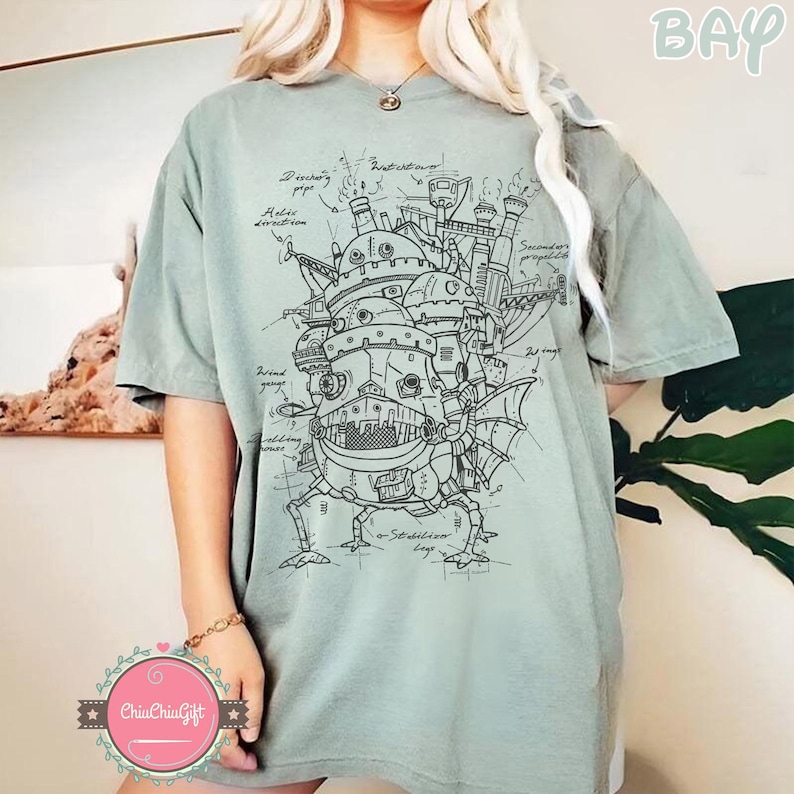 May include: Sage green t-shirt featuring a detailed black and white illustration of a fantastical, mechanical structure with architectural details and labeled components. The design is reminiscent of a blueprint or technical drawing.
