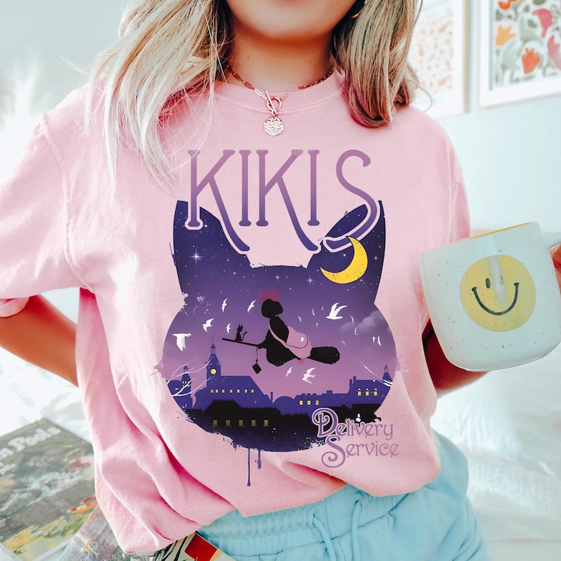 以下が含まれることがあります： 黒猫のシルエット、ほうきに乗った魔女、夜空のシーンが描かれた、明るいピンク色のスウェットシャツ。「KIKIS」の文字が猫の上に、「Delivery Service」の文字が下に書かれています。スマイリーフェイスのマグカップも写っています。