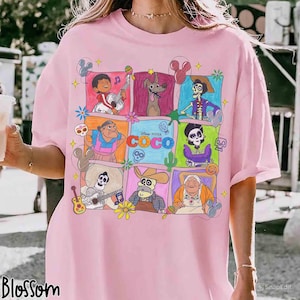 May include: Pink t-shirt featuring a colorful graphic of characters from the Disney Pixar movie Coco. The design includes various characters in square frames, with the movie's title prominently displayed. The shirt has short sleeves and a relaxed fit.