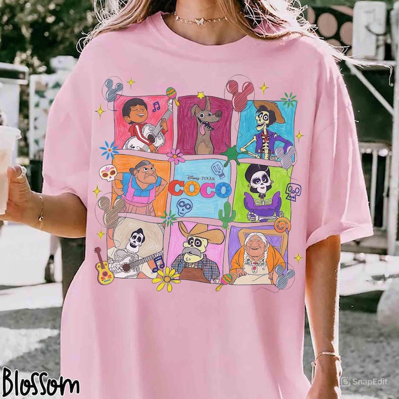 May include: Pink t-shirt featuring a colorful graphic of characters from the Disney Pixar movie Coco. The design includes various characters in square frames, with the movie's title prominently displayed. The shirt has short sleeves and a relaxed fit.