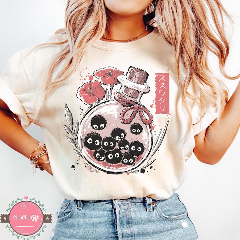 Puede incluir: Camiseta color crema con una ilustraci&oacute;n caprichosa de una botella de poci&oacute;n. Dentro de la botella hay varias criaturas negras y redondas con ojos grandes. Flores rojas y texto japon&eacute;s acent&uacute;an el dise&ntilde;o. La botella tiene un tap&oacute;n de corcho y est&aacute; atada con una cuerda roja.