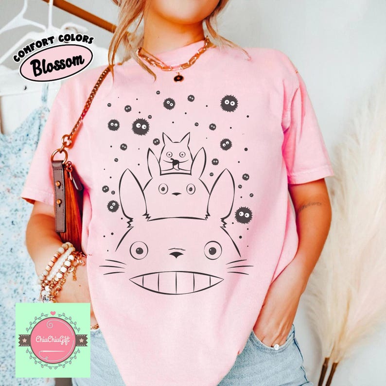 May include: A light pink t-shirt featuring a black and white illustration of three Totoro characters and several soot sprites. The shirt has the words "Comfort Colors" and "Blossom" printed on it.