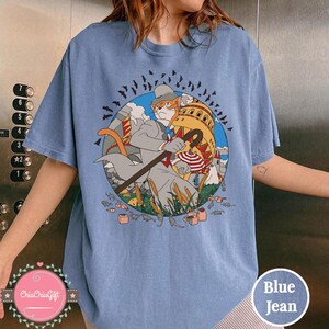 May include: Blue jean-colored t-shirt featuring a whimsical illustration of a cat dressed in a suit, holding a cane, and standing in front of a building. The design includes birds, clouds, and other cartoon elements. The shirt has a crew neck.