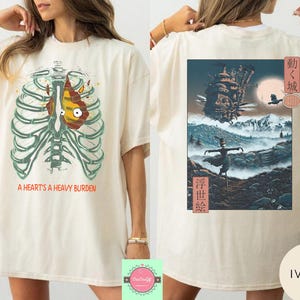 May include: Ivory-colored t-shirt featuring a graphic of a rib cage with a heart-shaped design and the text "A HEART'S A HEAVY BURDEN." The back of the shirt displays a colorful anime-style illustration with Japanese text.