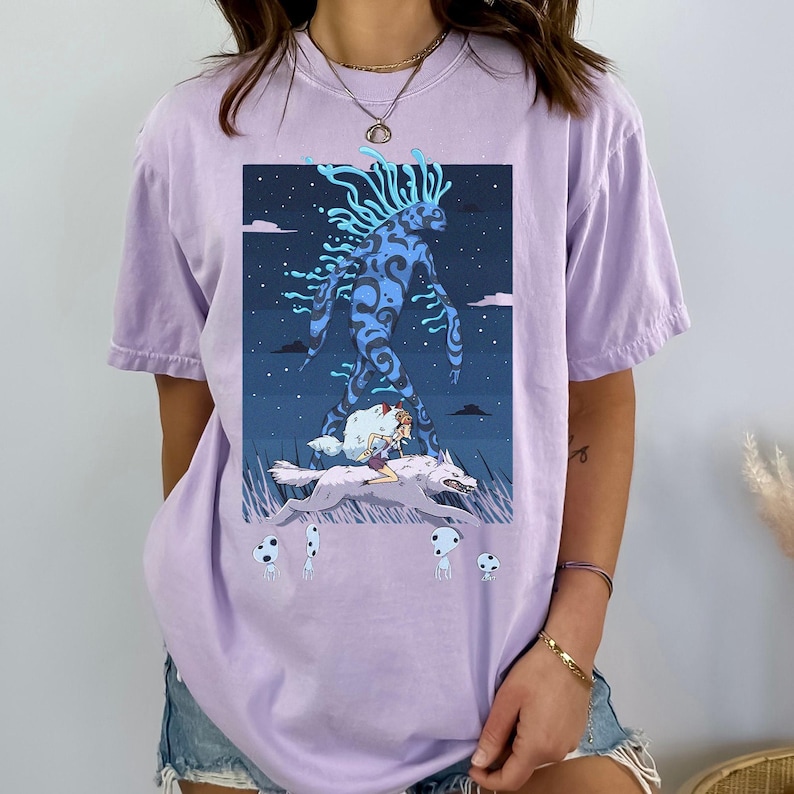 May include: A light purple t-shirt featuring a graphic illustration. The artwork depicts a blue, humanoid figure with swirling patterns, walking in a night scene. A person rides a wolf, and small mushroom-like figures are in the foreground.