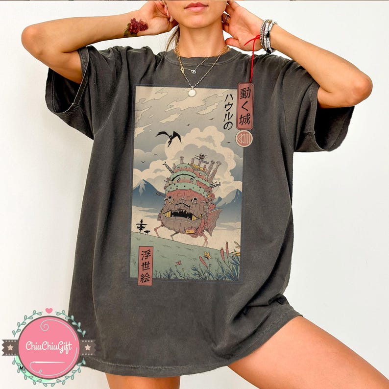 Pu&ograve; includere: T-shirt grigio scuro con un'illustrazione colorata di un castello in movimento e testo giapponese. Il design include un paesaggio montano, un uccello e caratteri giapponesi. La maglietta ha una vestibilit&agrave; ampia.