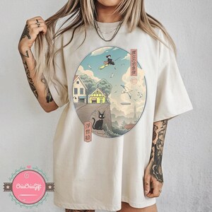 Op de afbeelding: Cr&egrave;mekleurig T-shirt met een cirkelvormige afbeelding met Japanse tekst en een landschapsillustratie. De illustratie toont een heks op een bezem, een zwarte kat, huizen en vliegende vogels. Het shirt heeft korte mouwen en een relaxte pasvorm.