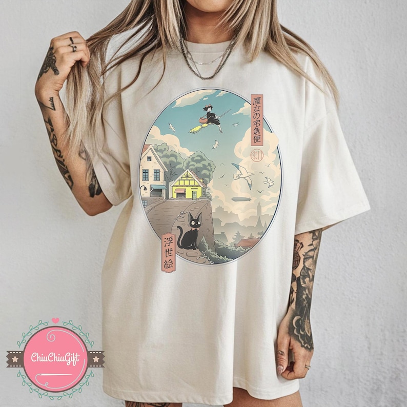 Op de afbeelding: Cr&egrave;mekleurig T-shirt met een cirkelvormige afbeelding met Japanse tekst en een landschapsillustratie. De illustratie toont een heks op een bezem, een zwarte kat, huizen en vliegende vogels. Het shirt heeft korte mouwen en een relaxte pasvorm.