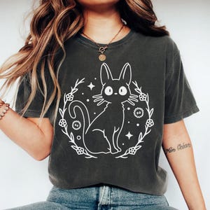 May include: Dark grey t-shirt featuring a white illustration of a cat inside a floral wreath with stars. The cat has large eyes and long ears. The shirt is a comfortable, casual style.