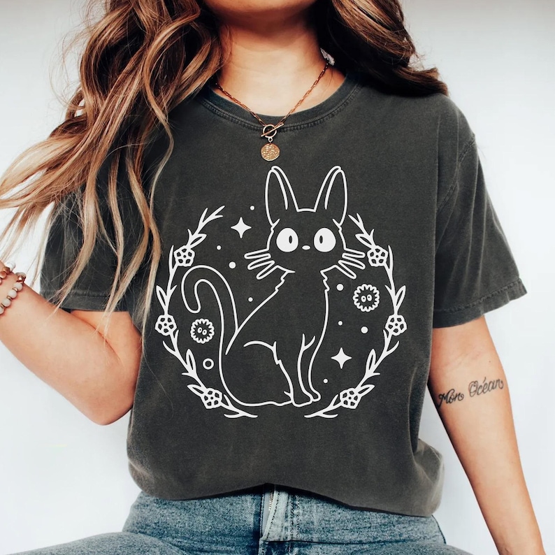 May include: Dark grey t-shirt featuring a white illustration of a cat inside a floral wreath with stars. The cat has large eyes and long ears. The shirt is a comfortable, casual style.