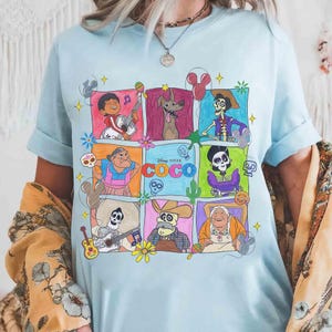 May include: Light blue t-shirt featuring a colorful graphic of characters from the Disney Pixar movie Coco. The design includes various characters in square frames with musical instruments and floral accents. The word "COCO" is also displayed.