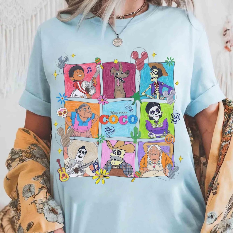May include: Light blue t-shirt featuring a colorful graphic of characters from the Disney Pixar movie Coco. The design includes various characters in square frames with musical instruments and floral accents. The word "COCO" is also displayed.