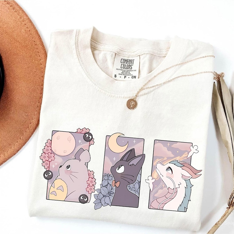 May include: Cream-colored Comfort Colors t-shirt featuring three framed anime-style illustrations. The illustrations depict a Totoro-like creature, a black cat, and a dragon, each gazing upwards. A gold necklace with a pendant rests on the shirt.