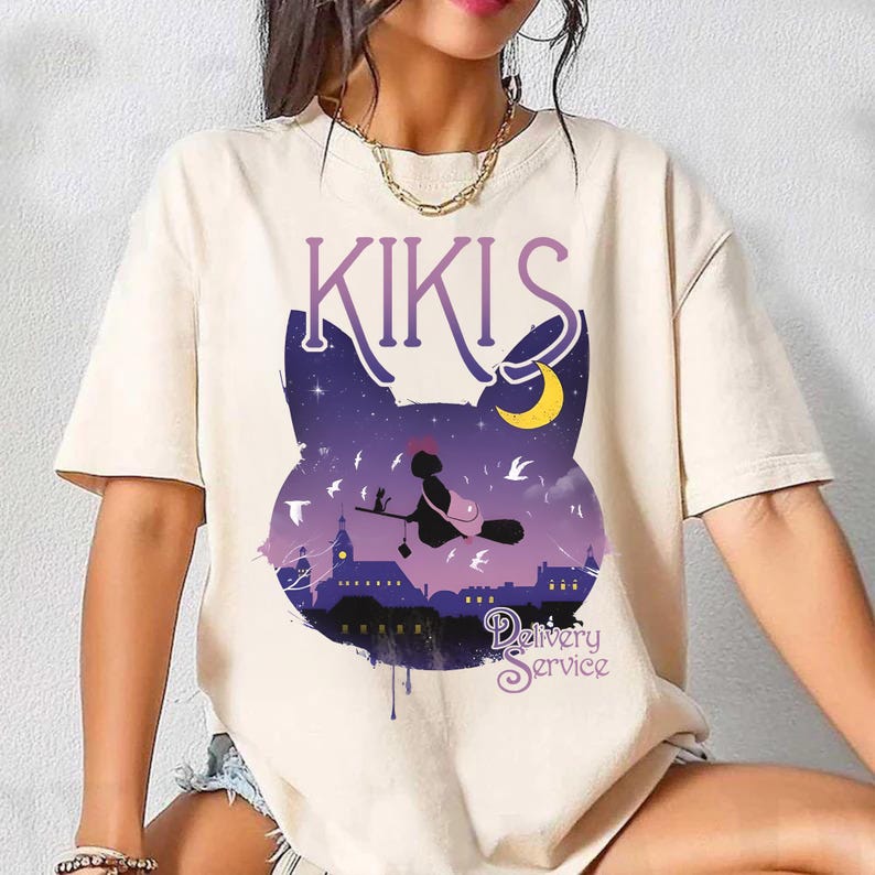 以下が含まれることがあります： クリーム色のTシャツ。紫色のグラフィックで、夜空のシーンと猫のシルエットが描かれています。「KIKIS」と「Delivery Service」の文字も含まれています。ほうきに乗った魔女も描かれています。