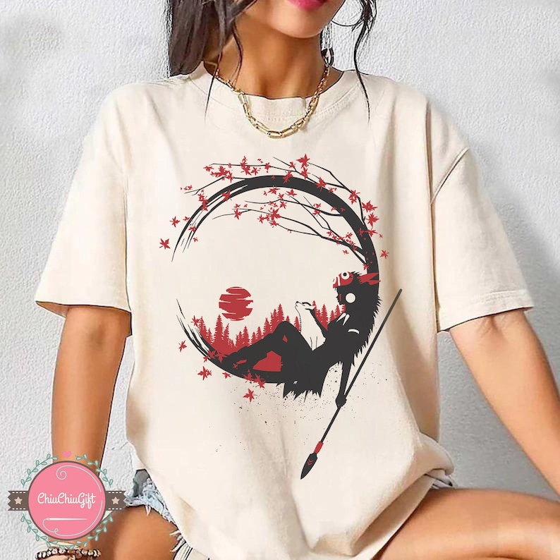 May include: Cream-colored t-shirt featuring a black and red graphic design. The design depicts a silhouette of a figure reclining within a circular frame, with a spear, a red sun, and a forest scene. The shirt has short sleeves.