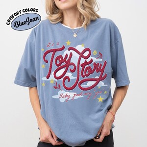 May include: A blue jean colored t-shirt featuring a graphic print with the red rope-style text "it's a Toy Story" and "Baby, just say yes". The design includes white clouds, yellow stars, and small red flowers. The shirt has a crew neckline.