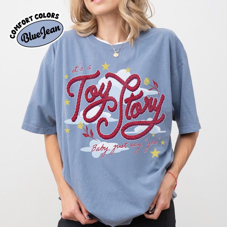 May include: A blue jean colored t-shirt featuring a graphic print with the red rope-style text "it's a Toy Story" and "Baby, just say yes". The design includes white clouds, yellow stars, and small red flowers. The shirt has a crew neckline.