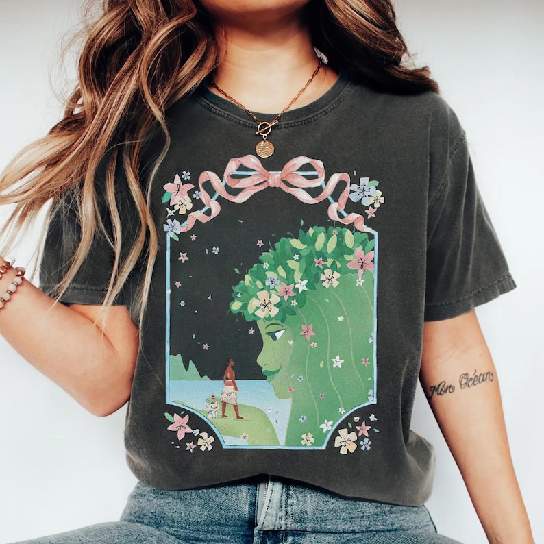 May include: Dark gray t-shirt featuring a graphic of a green-skinned woman with a floral crown and a pink ribbon. The design includes a landscape with a figure and a dog. The shirt has short sleeves.