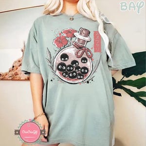 Puede incluir: Camiseta verde salvia con un gr&aacute;fico de una botella de vidrio con tap&oacute;n de corcho, flores rojas y criaturas negras y redondas con ojos. Texto japon&eacute;s impreso en el lado derecho de la botella.