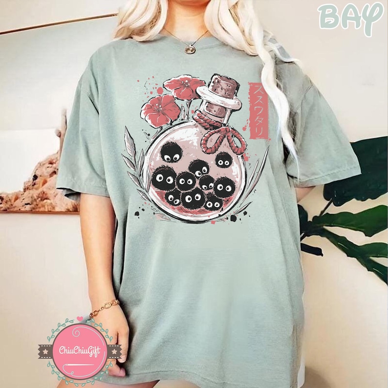 Puede incluir: Camiseta verde salvia con un gr&aacute;fico de una botella de vidrio con tap&oacute;n de corcho, flores rojas y criaturas negras y redondas con ojos. Texto japon&eacute;s impreso en el lado derecho de la botella.