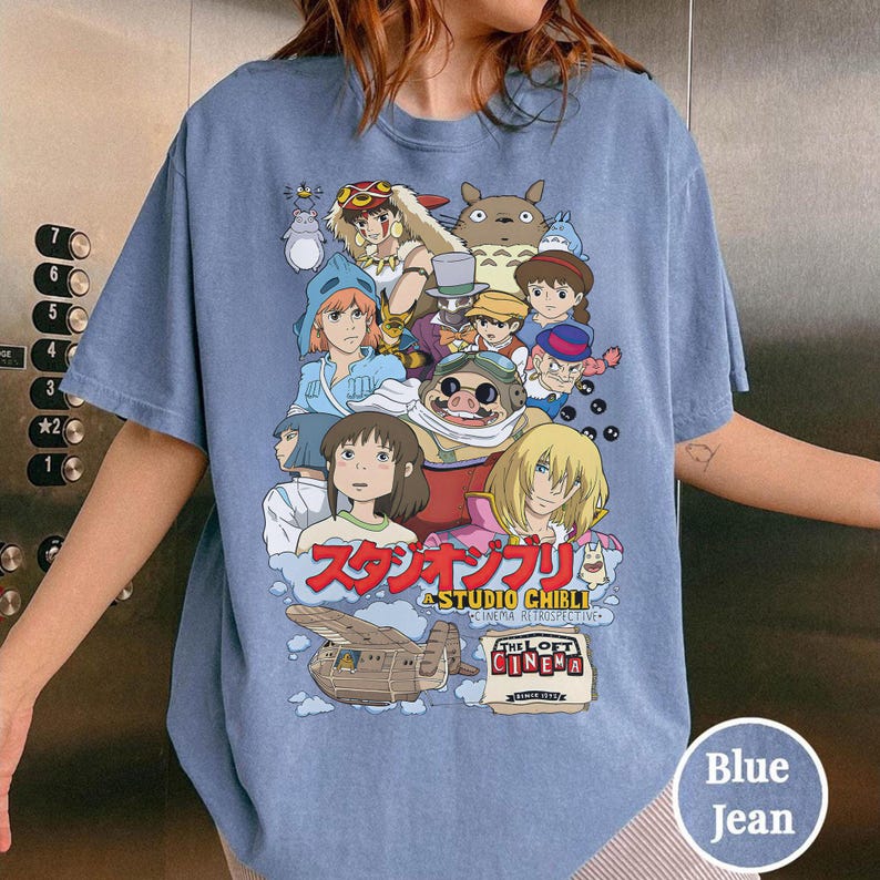 May include: Blue jean t-shirt featuring a colorful graphic of Studio Ghibli characters. The design includes characters from various animated films, with Japanese text and the words "THE LOFT CINEMA" and "STUDIO CHIBLI CINEMA RETROSPECTIVE".