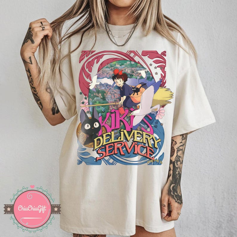 May include: A cream-coloured t-shirt with a vibrant graphic from the animated film Kiki's Delivery Service. The design features the text "Kiki's Delivery Service" alongside illustrations of Kiki, Jiji the cat, and a broomstick, set against a swirling blue and red backdrop.