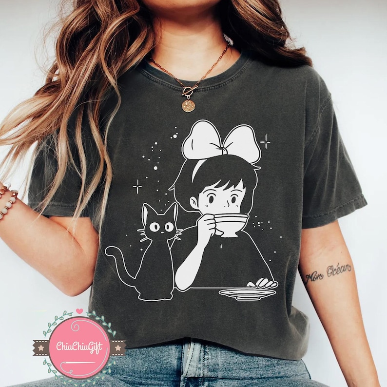 Pu&ograve; includere: T-shirt grigio scuro con un'illustrazione bianca in stile anime di una ragazza con un fiocco, che beve da una tazza, e un gatto nero. Il design include stelle e un piatto. Il testo "ChiaChiaGift" &egrave; visibile.