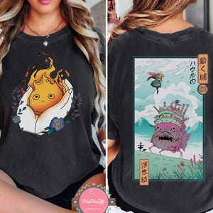 K&ouml;nnte beinhalten: Dunkelgraues T-Shirt mit einem Front-Grafik eines Cartoon-Feuergeistes, der in H&auml;nden gehalten wird, mit Lavendel und blauen Blumen. Die R&uuml;ckseite des Shirts hat eine rechteckige Grafik mit japanischem Text und einer Schlossillustration.