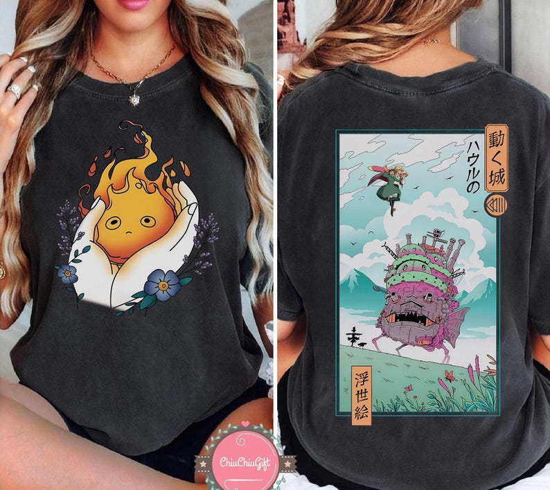 K&ouml;nnte beinhalten: Dunkelgraues T-Shirt mit einem Front-Grafik eines Cartoon-Feuergeistes, der in H&auml;nden gehalten wird, mit Lavendel und blauen Blumen. Die R&uuml;ckseite des Shirts hat eine rechteckige Grafik mit japanischem Text und einer Schlossillustration.