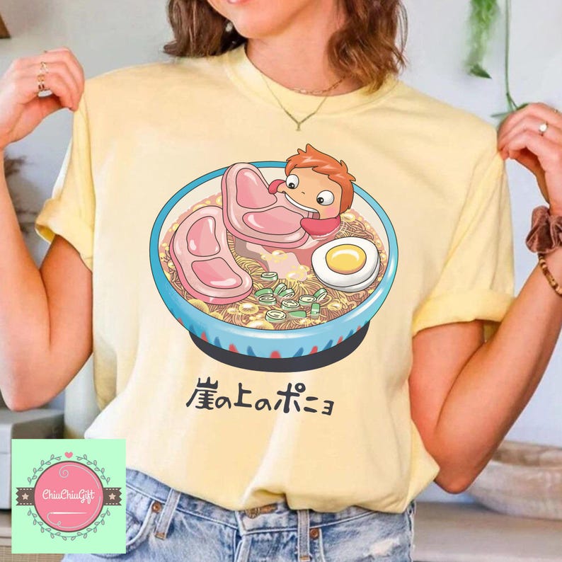 May include: Yellow t-shirt featuring a cartoon illustration of a bowl of ramen with a character from the animated film Ponyo. The bowl has a blue rim and contains noodles, meat, and an egg. Japanese text is below the bowl.