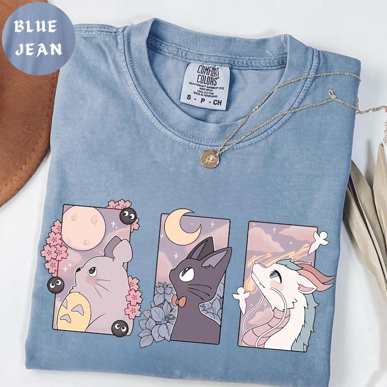 May include: A light blue Comfort Colors t-shirt featuring three anime-style illustrations. The illustrations include a gray creature with a pink flower, a black cat with a bow tie, and a white dragon. The shirt also has the text "BLUE JEAN".