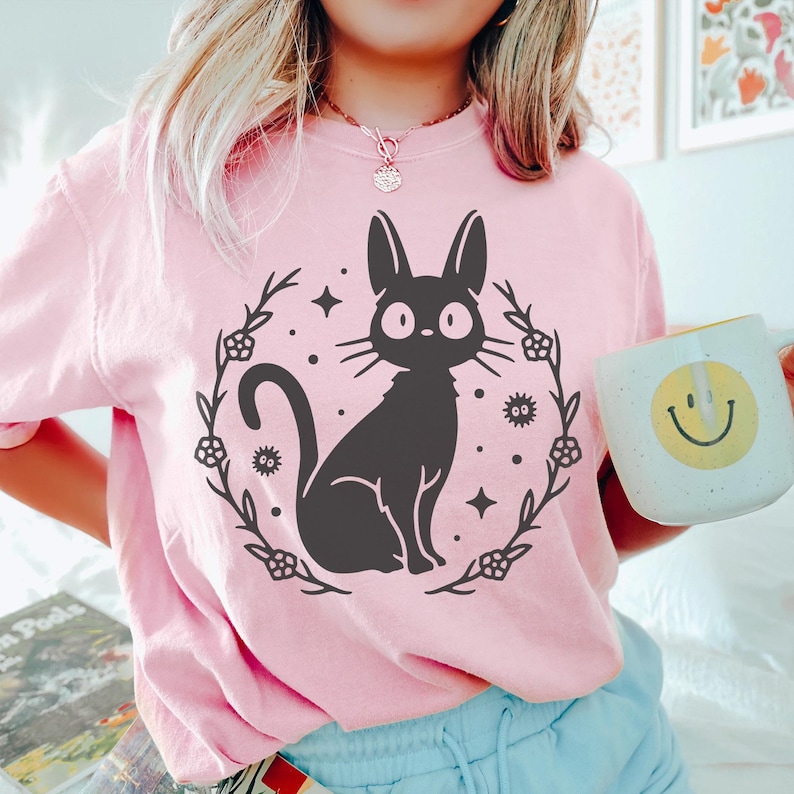 May include: A light pink sweatshirt featuring a black cat illustration within a floral wreath, surrounded by stars and dots. The cat has large, round eyes. A white mug with a yellow smiley face is held in the hand.