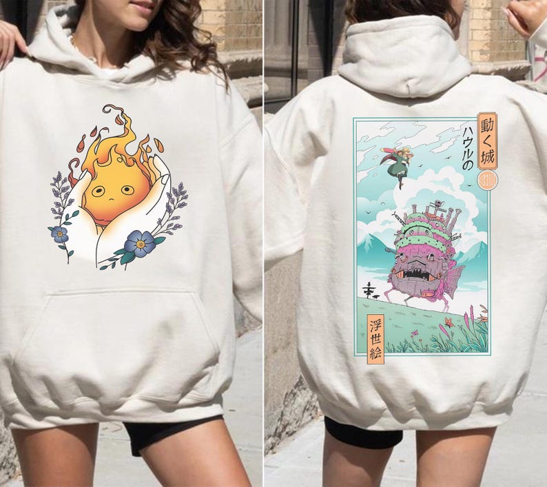 K&ouml;nnte beinhalten: Cremefarbener Kapuzenpullover mit einem Front-Design eines Cartoon-Feuergeistes in einer Hand, umgeben von floralen Elementen. Auf der R&uuml;ckseite befindet sich eine farbenfrohe Illustration eines sich bewegenden Schlosses mit japanischem Text. Ein l&auml;ssiges Kleidungsst&uuml;ck.