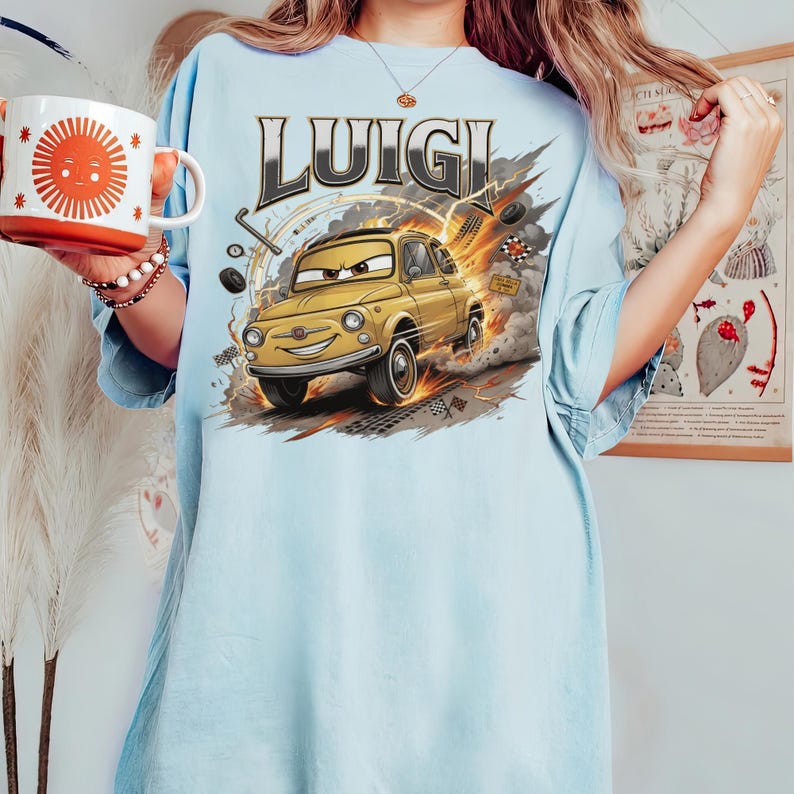 Pode incluir: Uma t-shirt azul-clara com uma estampa gr&aacute;fica da personagem Luigi do filme Carros. O design mostra um carro vintage amarelo com um rosto sorridente, rodeado por elementos de corrida, fumo e o texto LUIGI em letras a negrito.