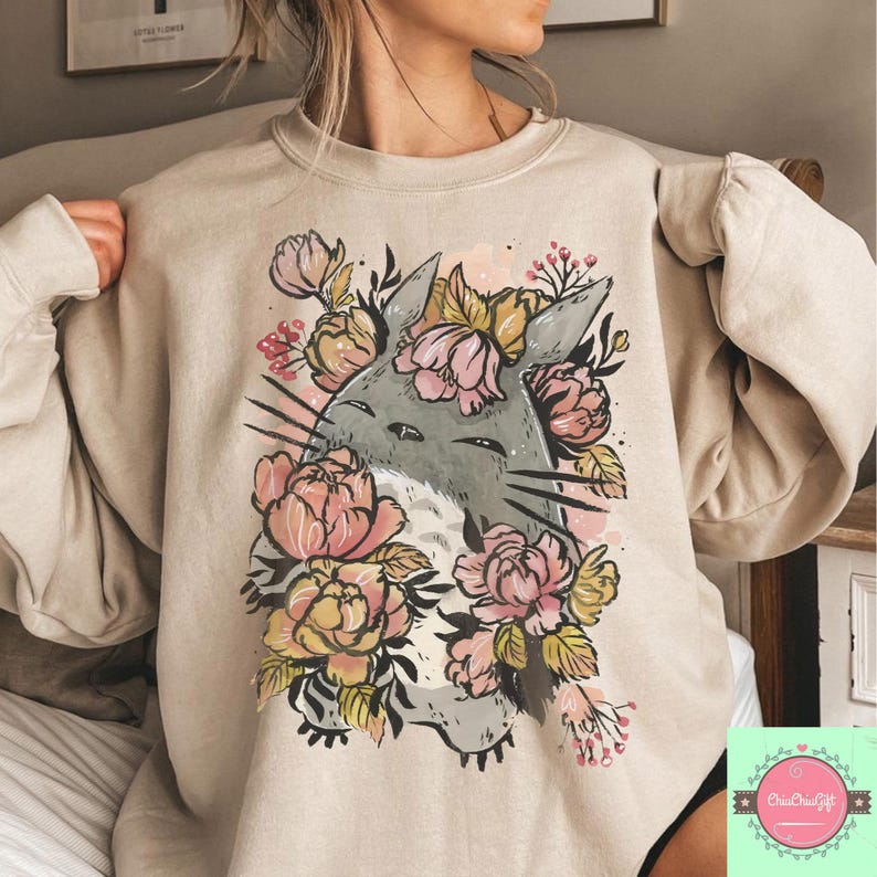 May include: Beige sweatshirt featuring a gray cartoon character surrounded by pink and yellow floral elements. The character has long whiskers and pointed ears. The design is centered on the front of the sweatshirt.