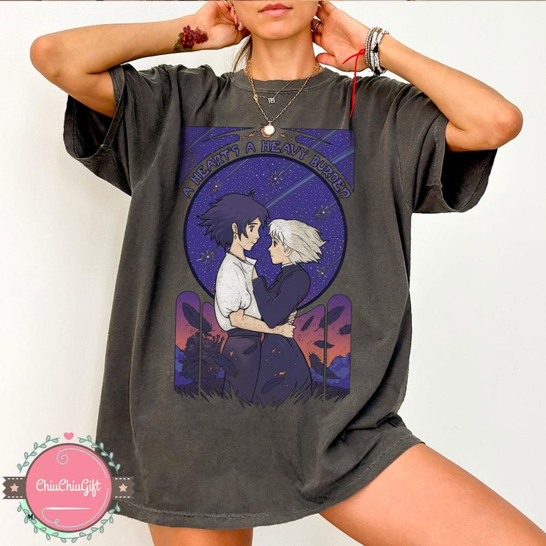 Puede incluir: Camiseta gris oscuro con una ilustraci&oacute;n de estilo anime de dos personajes abraz&aacute;ndose bajo un cielo estrellado. El dise&ntilde;o incluye el texto "A Heart's A Heavy Burden" en un marco circular. La camiseta tiene un corte holgado.