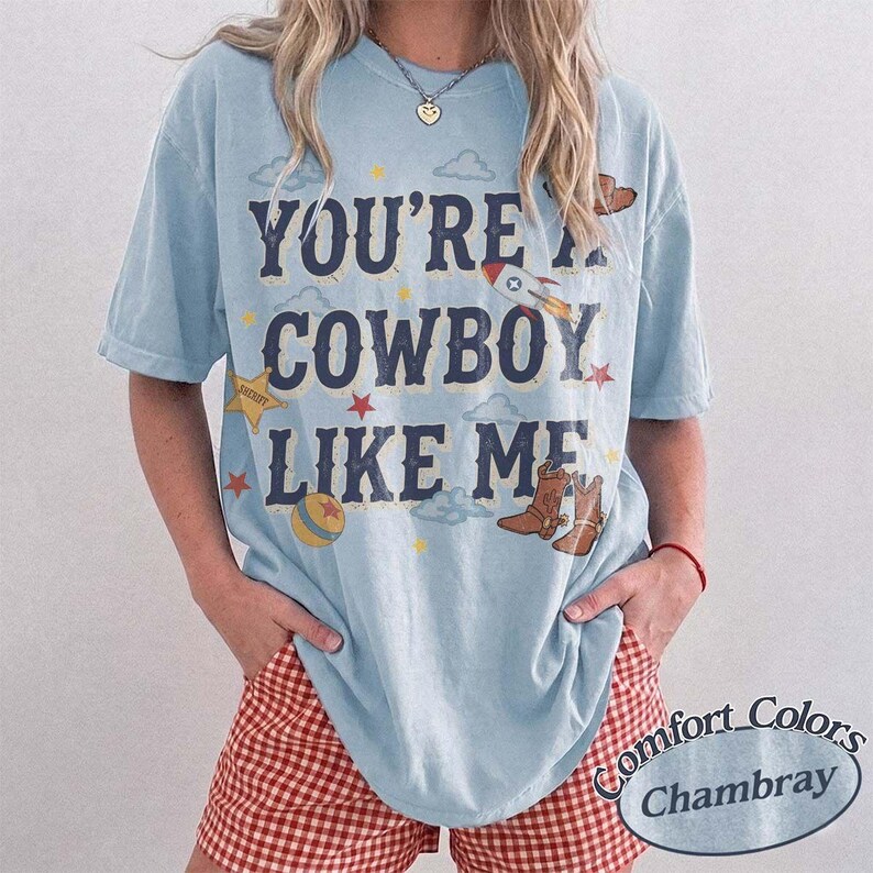 May include: A light blue t-shirt featuring the dark blue text "YOU'RE A COWBOY LIKE ME" printed on the front. The design includes small illustrations of a rocket, a sheriff star, a ball, and brown cowboy boots scattered among clouds and stars.